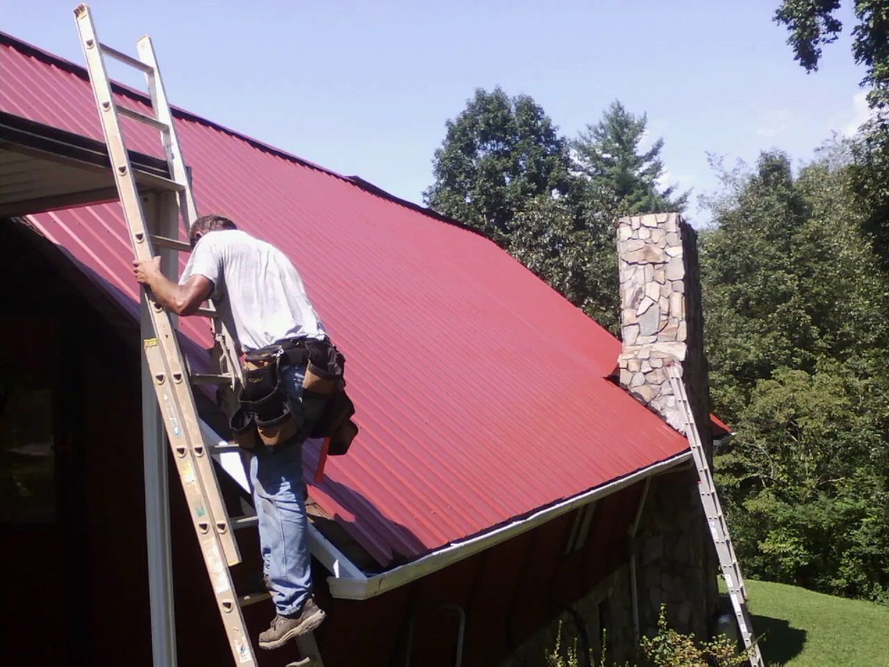 Roofer on a ladder beside a red metal roof and stone chimney — Soffit & Fascia Repair by Roofing Brothers in Picabo
